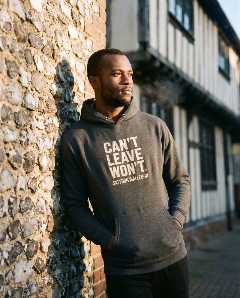 Man in the charcoal CAN'T LEAVE. WON'T. hoodie leaning on an old flint wall, timber-framed building behind